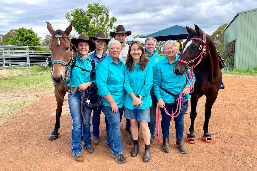 Men and women in aqua green shirts smile next to two horses.