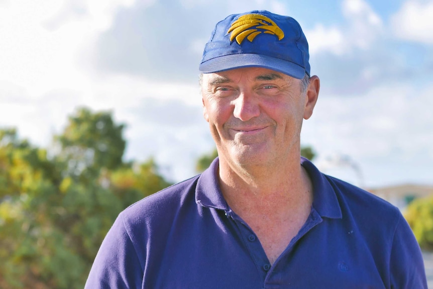 A man wearing a blue baseball cap and blue polo shirt grins as he stands outdoors.