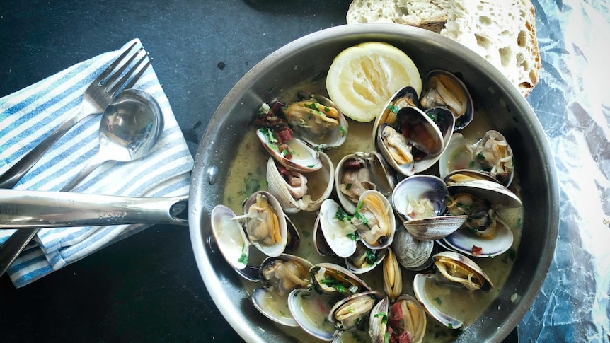 A skillet full of cooked mussels in a creamy sauce sits on a table next to a hunk of rustic bread.