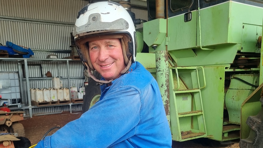 A man is wearing up a helmet while holding the handlebars of a motorbike. He's standing in a shed around farm machinery