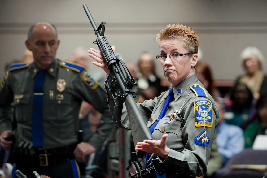 A woman in a police uniform holds up an assault style rifle in a courtroom.