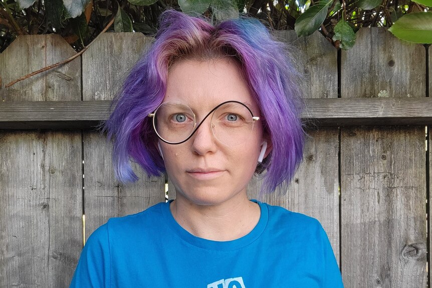 a person with purple hair and hip glasses stands in a backyard looking at the camera. 