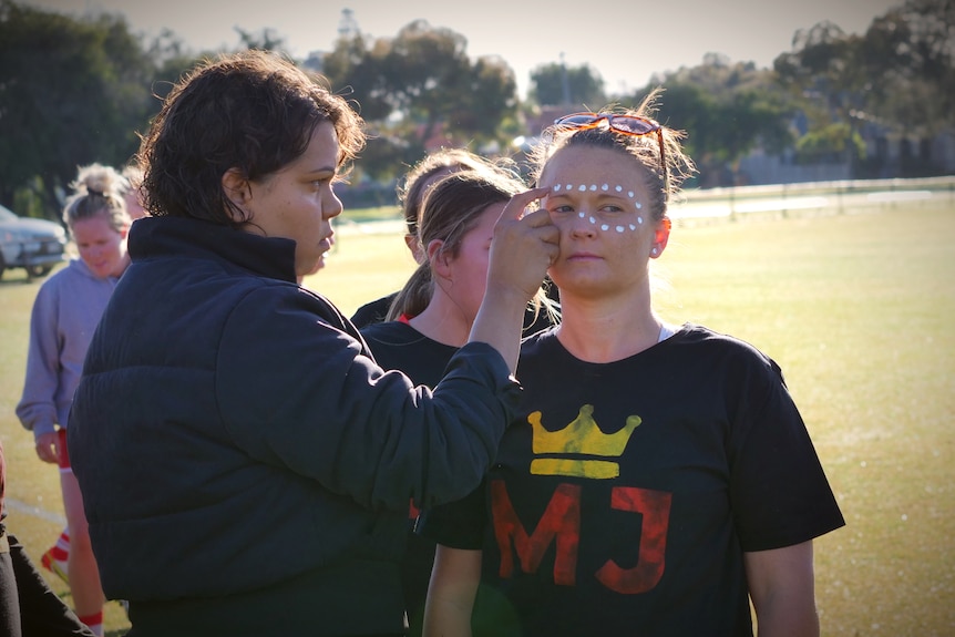 A woman applies white dot paint to another woman's face