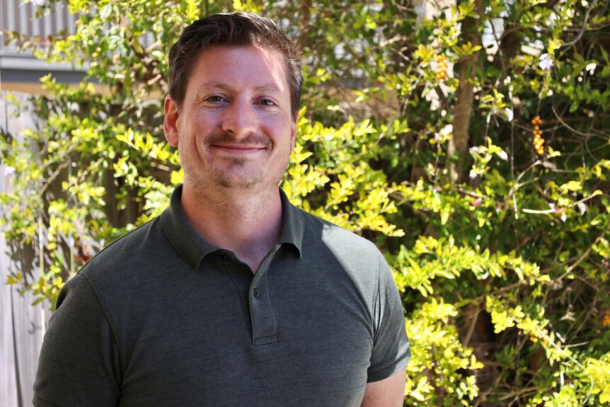 A man in a khaki shirt smiling in front of a tree