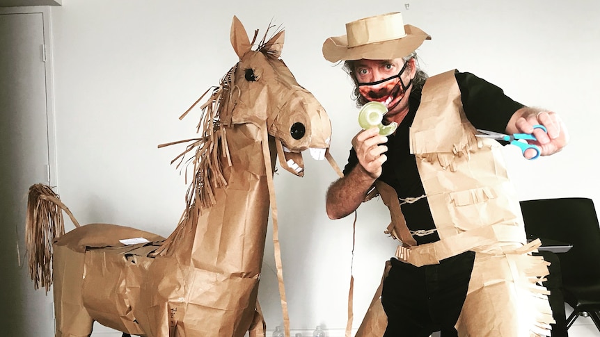 A man in a hotel room dressed as a cowboy standing next to a horse made from various objects.