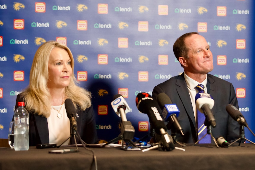 A woman and a man in business attire are both sitting behind microphones at a press conference