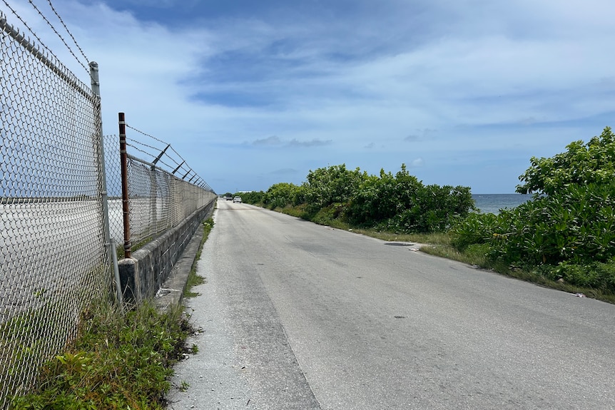 A road with a fence running along one side and the ocean on the other side, blue skies.