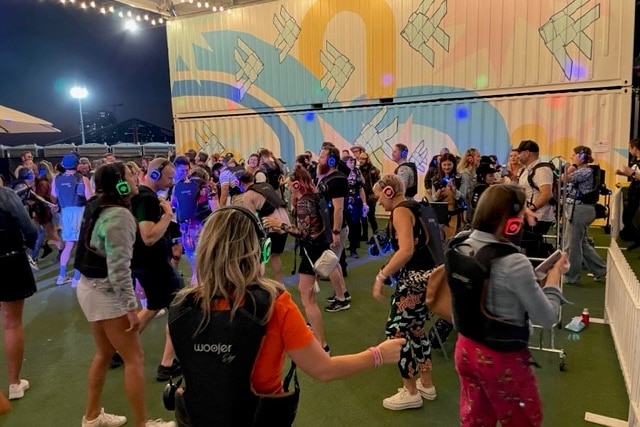 A group of people dance at a semi-outdoor festival, wearing headphones and futuristic-looking vests.