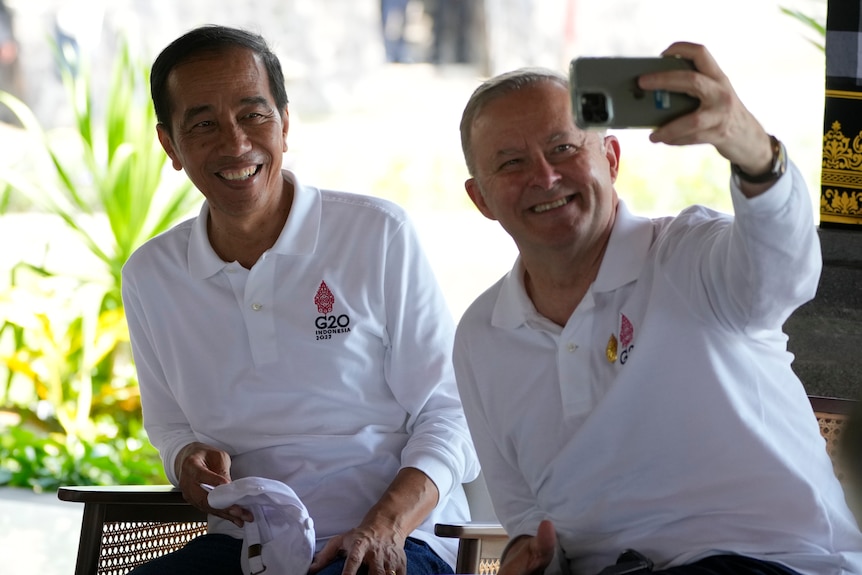 Three smiling men in white tops sit together as one takes a selfie with a moblie.