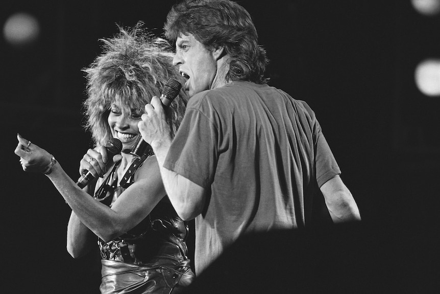 A black-and-white poto of a smiling Tina Turner and grimacing Mick Jagger singing together.