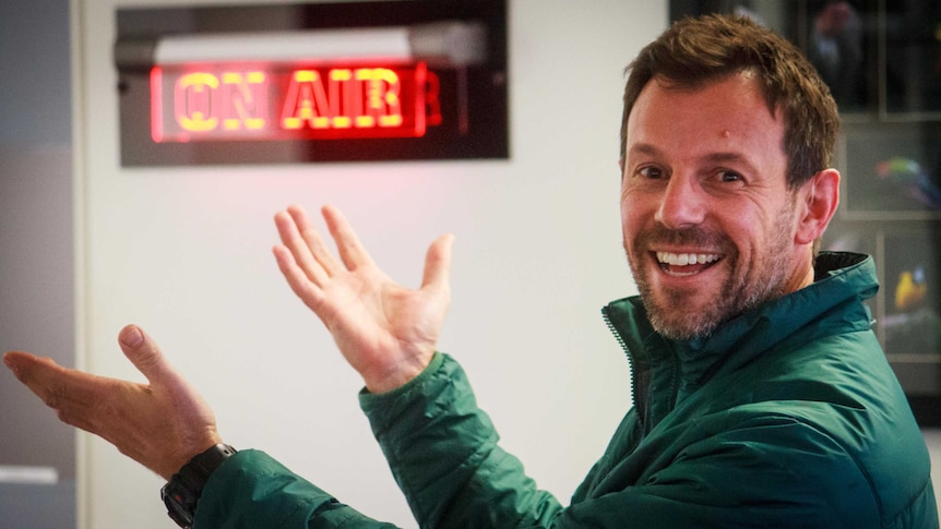 A man in radio studio with his hands raised smiling at the camera.