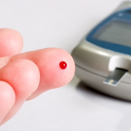 A fingertip with a spot of blood on it, with the machine used to measure glucose in the background.
