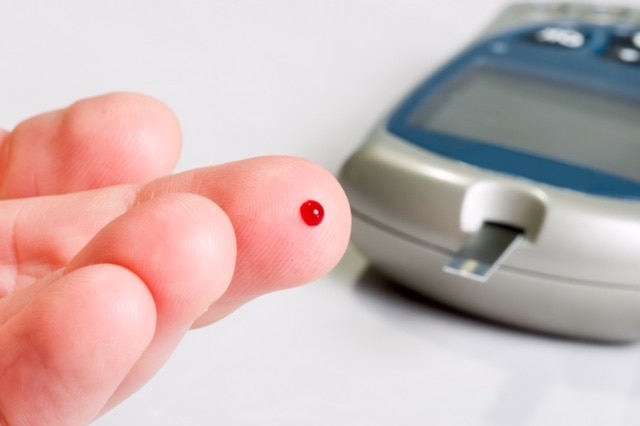 A fingertip with a spot of blood on it, with the machine used to measure glucose in the background.