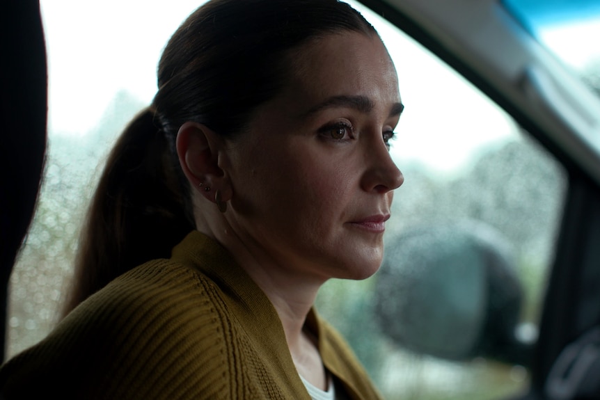 A woman sits in the passenger seat of a car, looking to one side with a serious expression. The windows have rain drops on them.