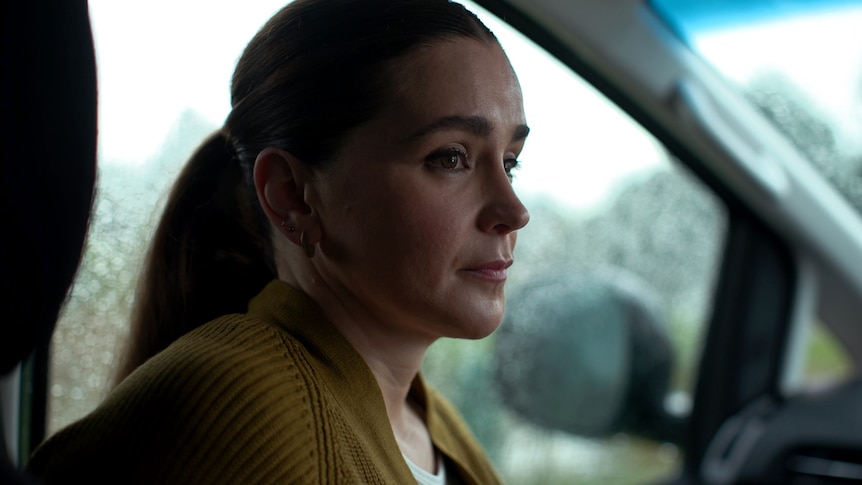 A woman sits in the passenger seat of a car, looking to one side with a serious expression. The windows have rain drops on them.