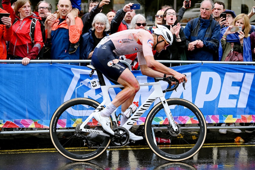 A Dutch cyclist rides past a row of fans in the wet with blood and bruises visible on his leg. 
