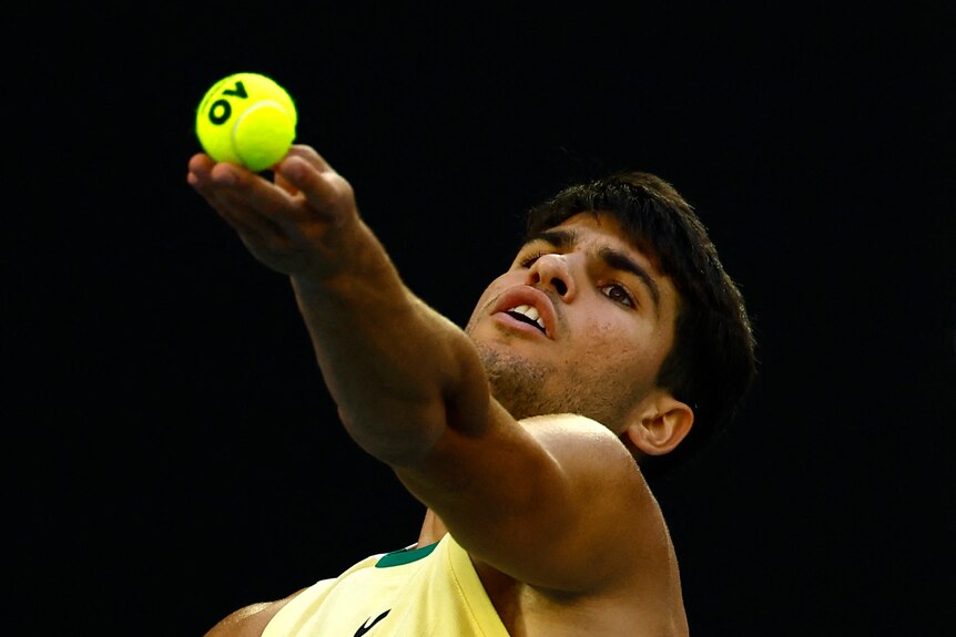Male tennis player is about to release the ball for a serve, looking skyward in a yellow top