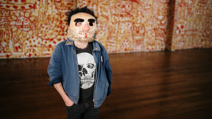 a man wearing a puppet-like mask with dark eyebrows and hair stands in a room with a mural behind him