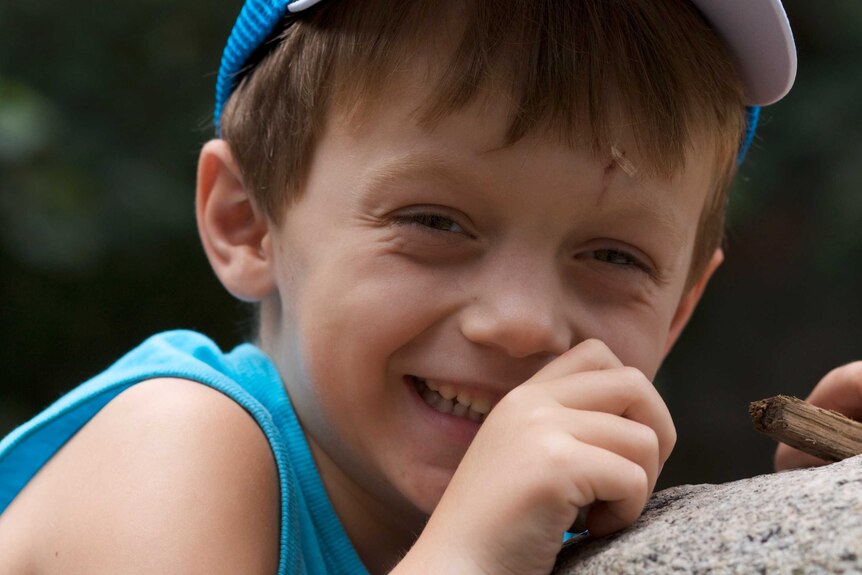 A little boy smiling big while wearing a hat and covering his mouth slightly