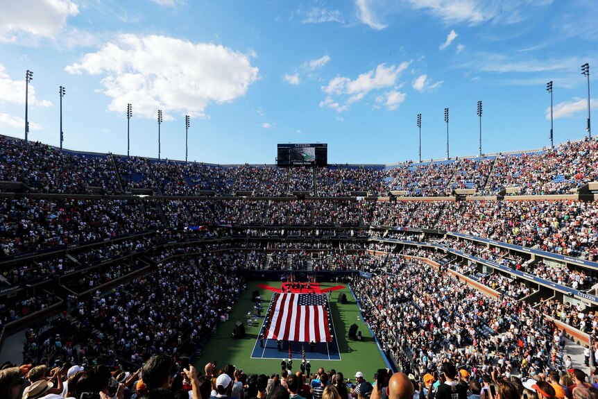 View of a packed tennis grandstand with a big American flag spread out over the tennis court below.