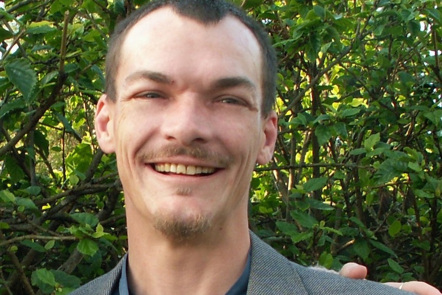 Alan Taylor poses for a picture wearing a blue shirt and grey jacket with a smile on his face.