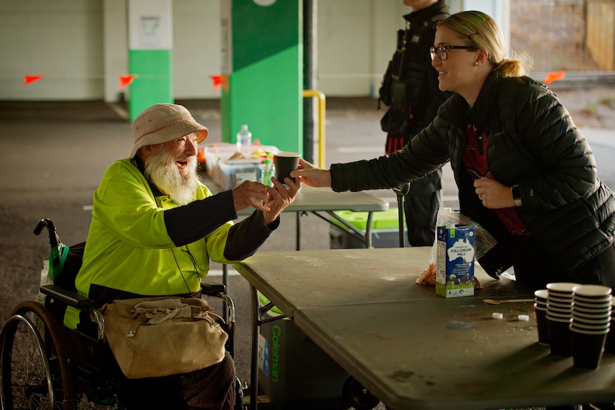 A woman wearing a black jacket serves a hot coffee to a man in wheelchair. They are both happy and smiling at each other.