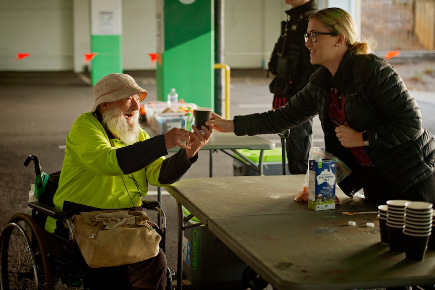 Une femme portant une veste noire sert un café chaud à un homme en fauteuil roulant.  Ils sont tous les deux heureux et souriants l'un à l'autre.