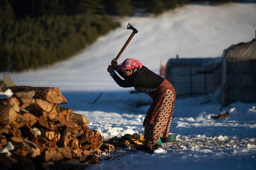 A woman cuts firewood outside in the snow.