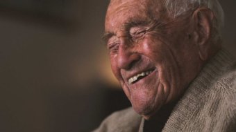 A very old man in a tan cardigan laughs against a black background.