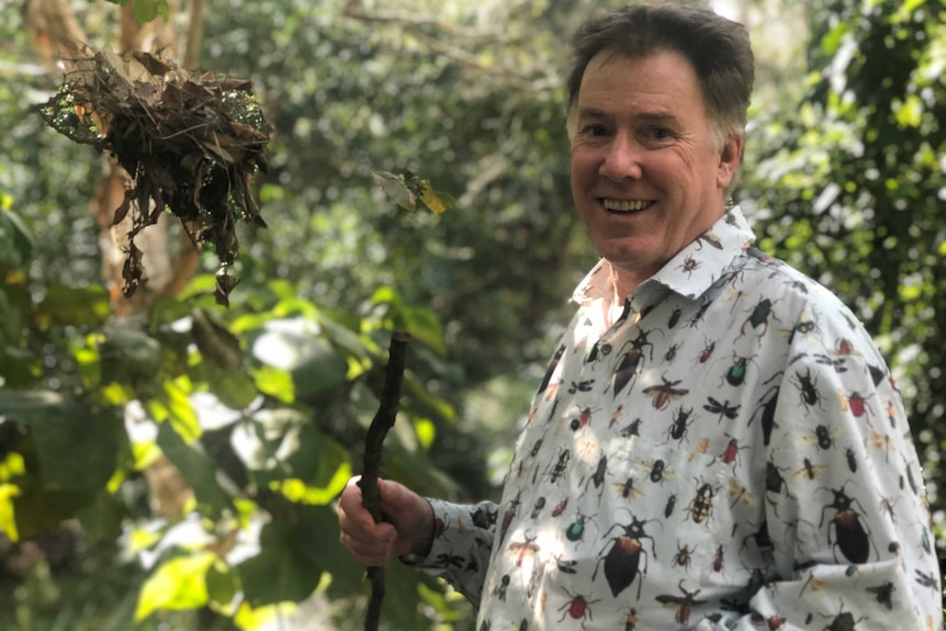 A man with short hair, wearing a bug-patterned shirt, holds a stick while standing in the bush.