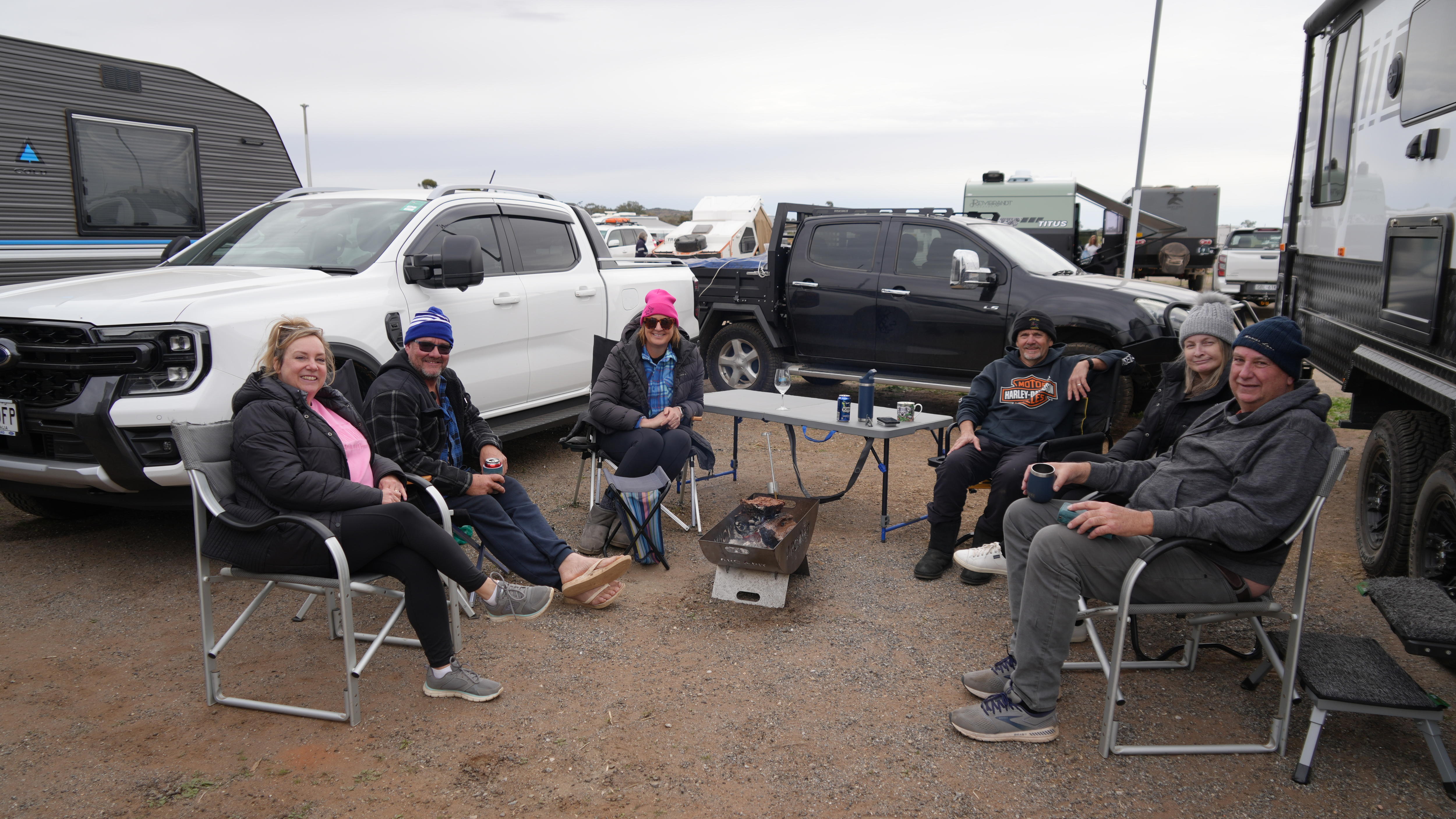 Group of six people sit around campfire on fold up chairs facing camera, next to a table, utes and a caravan