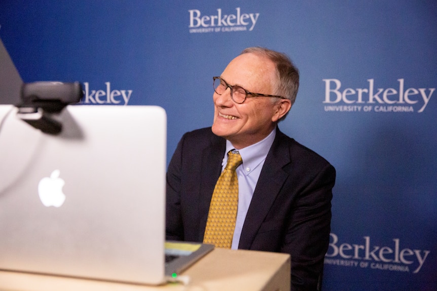 A man smiles at his laptop as he gives an interview in front of a press wall that says Berkeley.