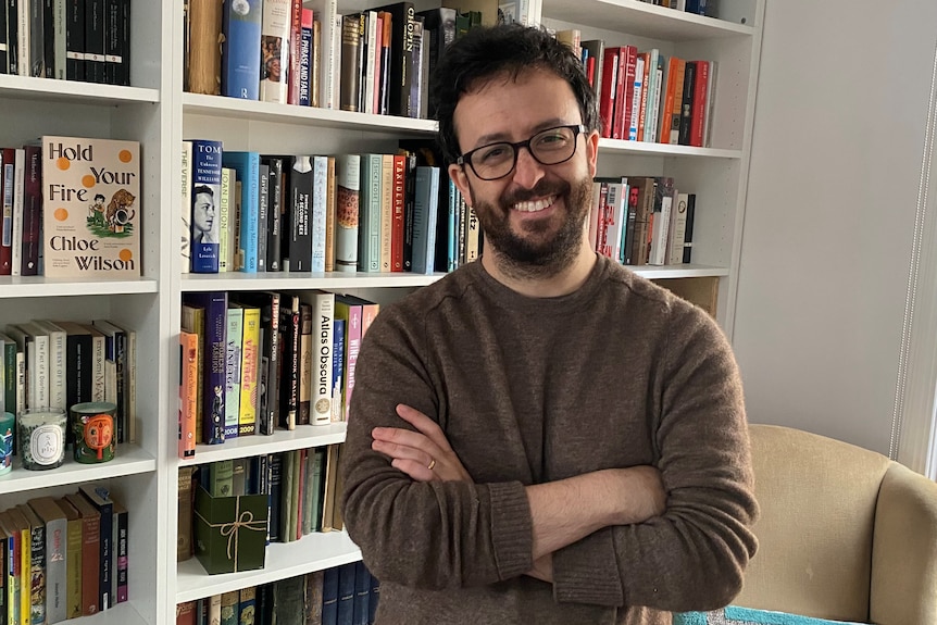 a smiling man in a brown jumper against a bookcase