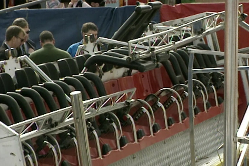 Four men huddle at the back of a broken amusement ride with several harnesses open