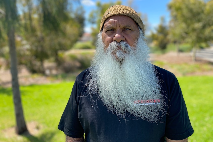 A senior Aboriginal man with a long grey beard stands outdoors. His expression is serious.
