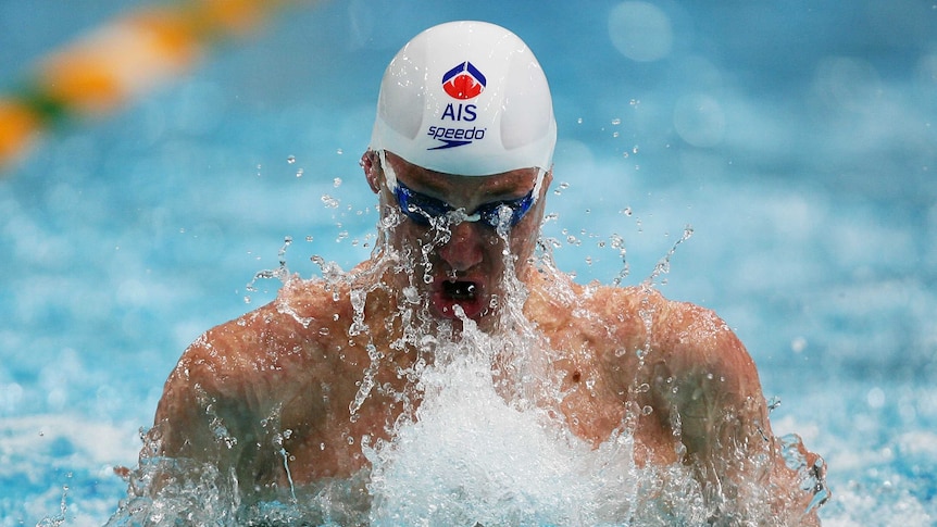 A male swimmer in the pool mid-stroke, clearly wearing an Australian Institute of Sport (AIS) swimming cap