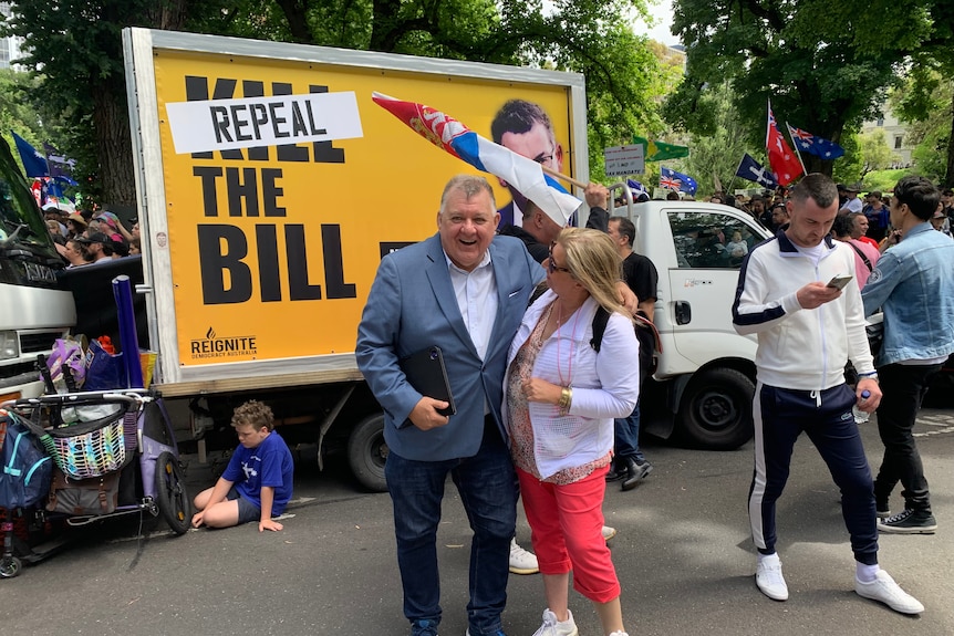 MP Craig Kelly smiles in front of a billboard saying 'repeal the bill'.