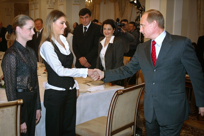 Vladimir Putin shaking hands with a young woman with long blonde hair while the people around them smile or look nervous