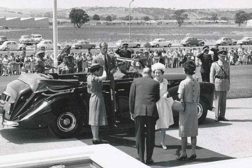Black and white photo of the Queen, smiling and shaking hands with a man, standing near a car, with a crowd of people behind her.