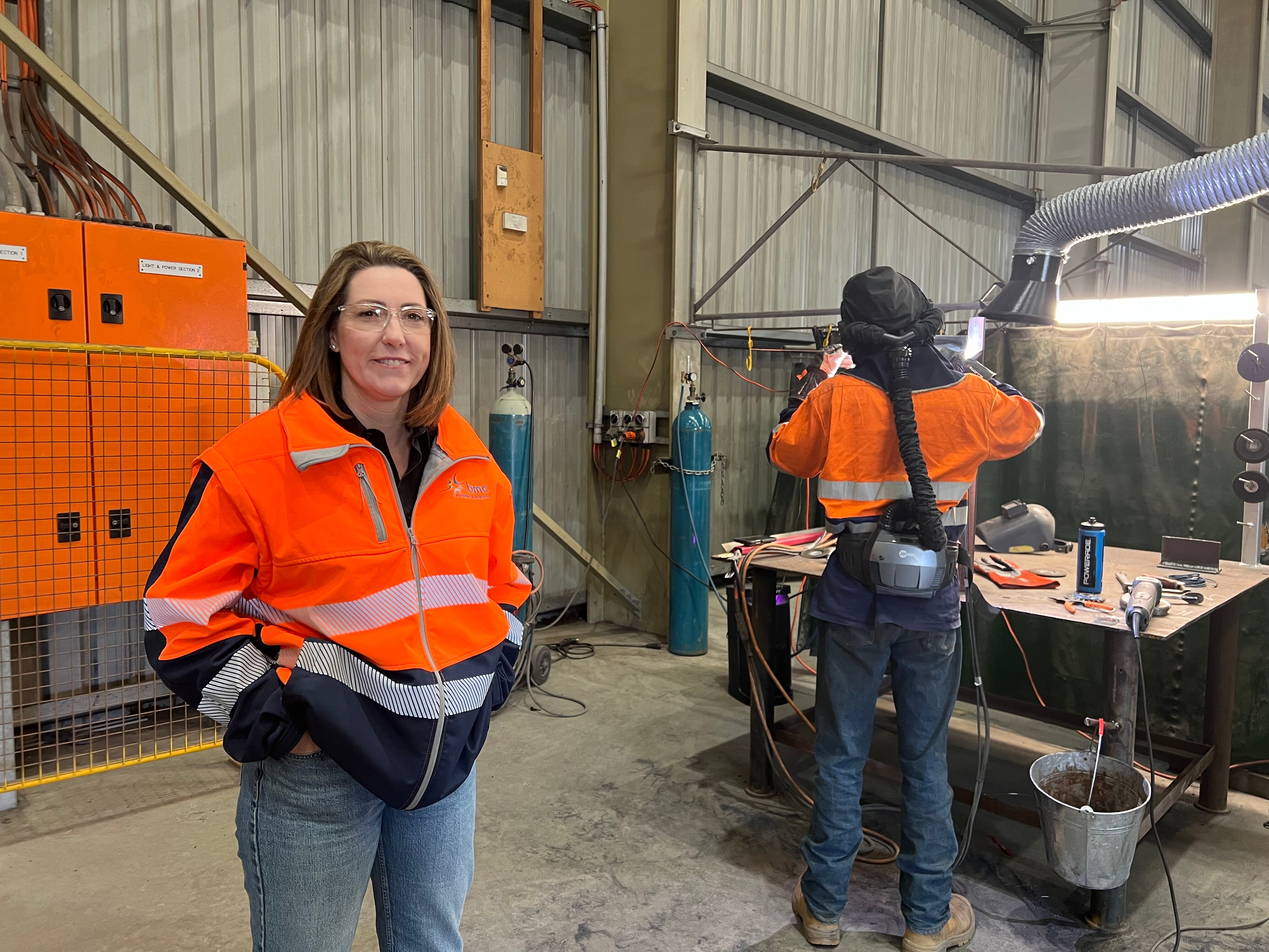 Woman standing in high vis jacket infront of a tradie working in a manufacturing workshop with various tools infront of him.