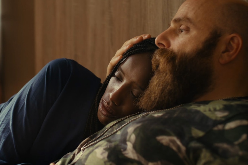 An African American woman rests her head on the chest of a white man with a beard, as they lay in bed together.