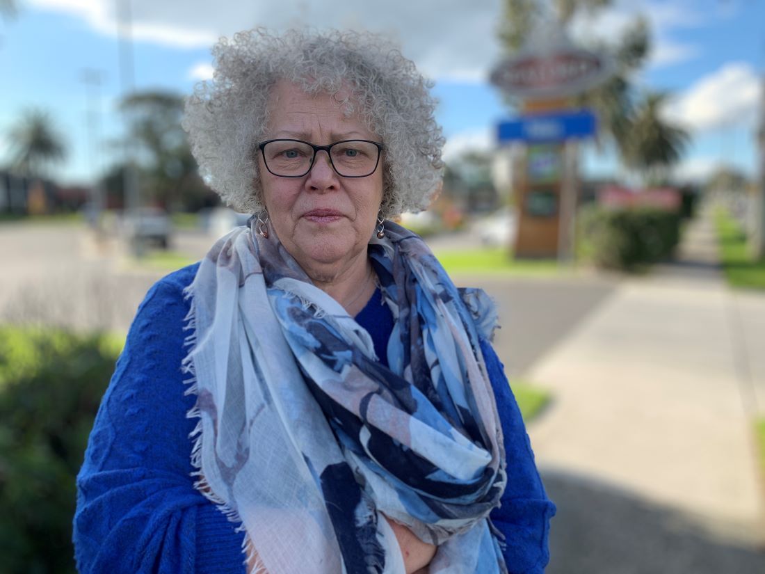 A woman with grey curly hair and a blue and grey scarf stands on a footpath.