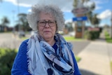 A woman with grey curly hair and a blue and grey scarf stands on a footpath.