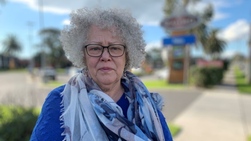 A woman with grey curly hair and a blue and grey scarf stands on a footpath.