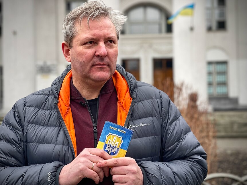 A man wearing a black puffer jacket with orange lining clutches a yellow and blue Ukrainian document, standing outside