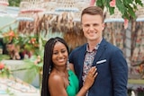 A man in a suit and a woman in a green dress smile at the camera with a tropical background behind.