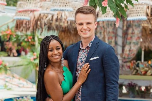 A man in a suit and a woman in a green dress are smiling at the camera with a tropical background behind.