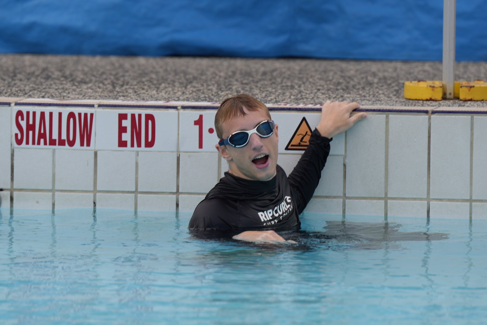 A man wearing goggles and a long sleeved swimming shirt holds onto the wall while in a swimming pool.