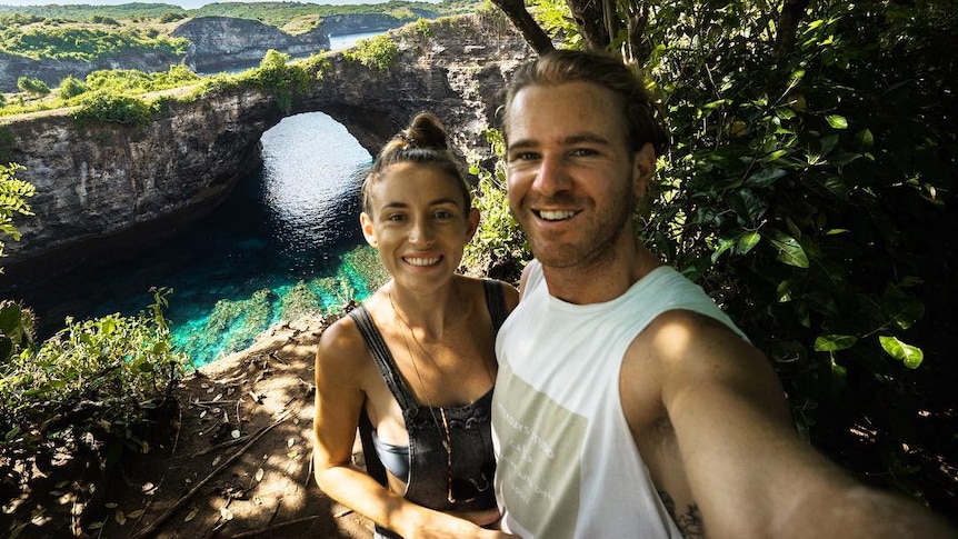 Jolie King and Mark Firkin take a selfie together with water and cliffs in the background.
