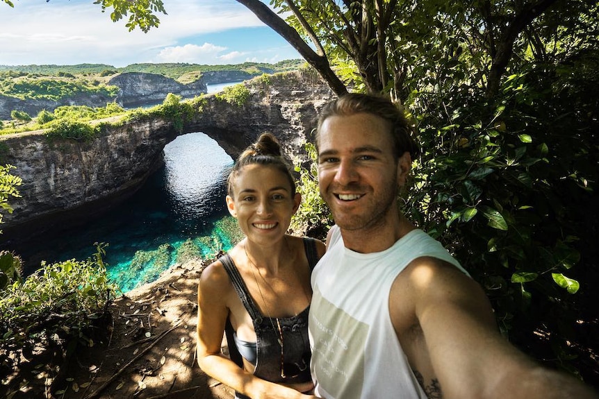 Jolie King and Mark Firkin take a selfie together with water and cliffs in the background.